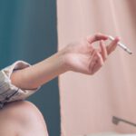 Risk Tool - Unrecognizable female sitting at white table with typewriter near blue and pink curtain while holding cigarette in hand