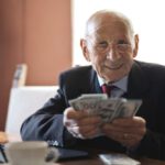 Retirement Income - Confident senior businessman holding money in hands while sitting at table near laptop