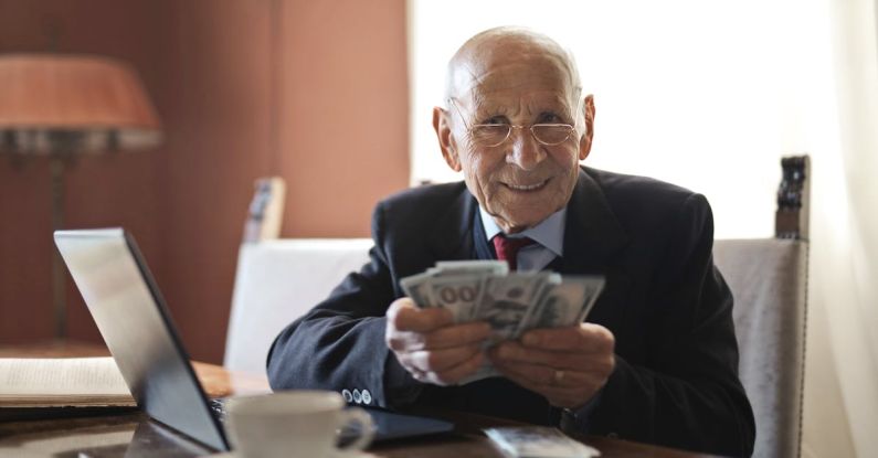 Retirement Income - Confident senior businessman holding money in hands while sitting at table near laptop