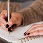 529 Plan - Crop unrecognizable female with stylish manicure sitting at black desk with keyboard and smartphone and taking notes with silver pen in notepad