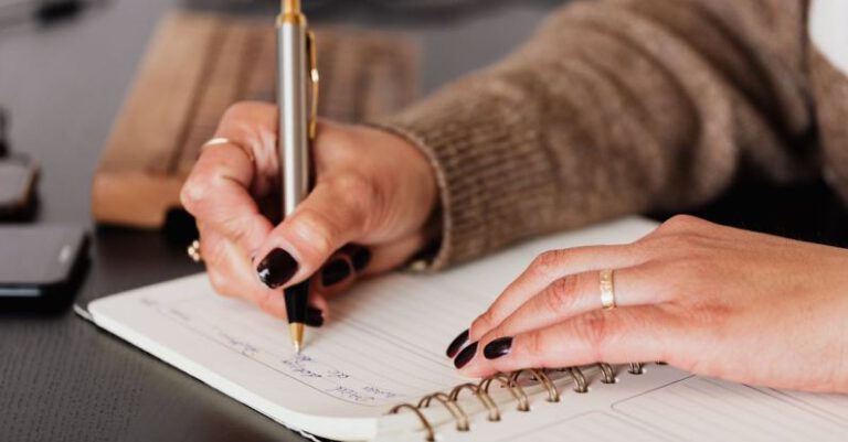529 Plan - Crop unrecognizable female with stylish manicure sitting at black desk with keyboard and smartphone and taking notes with silver pen in notepad