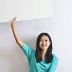 International Real Estate - Cheerful Asian woman sitting cross legged on floor against white wall in empty apartment and showing white blank banner