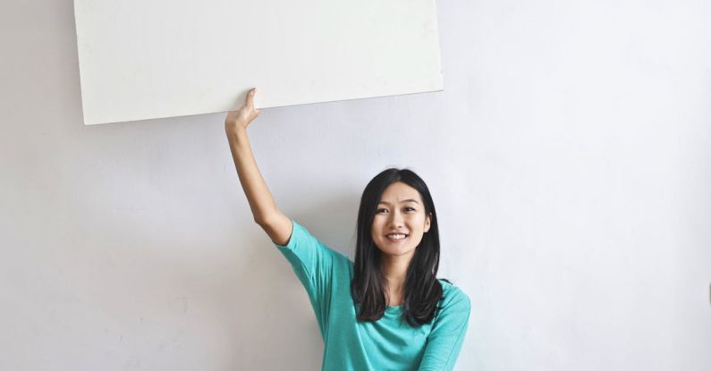 International Real Estate - Cheerful Asian woman sitting cross legged on floor against white wall in empty apartment and showing white blank banner