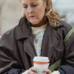 Urbanization - Lonely elderly female in coat with hot drink to go sitting with crossed legs on bench while looking down
