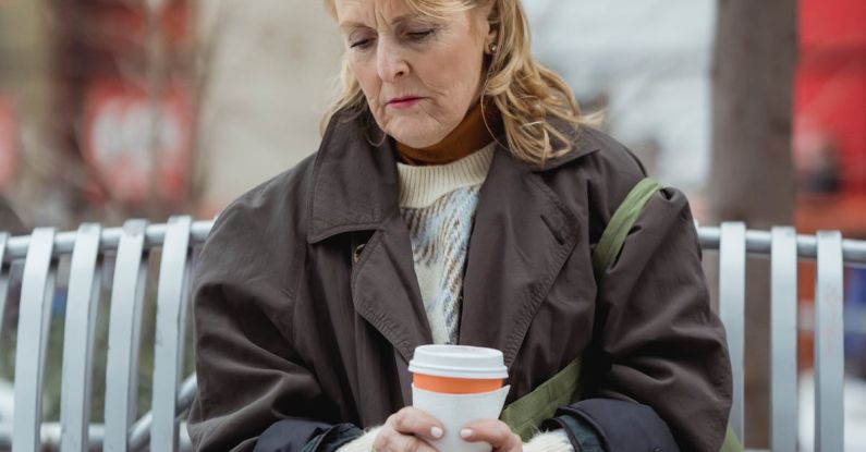 Urbanization - Lonely elderly female in coat with hot drink to go sitting with crossed legs on bench while looking down